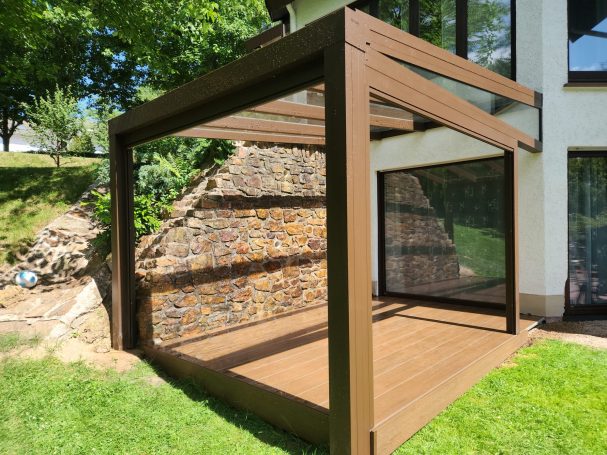 Moderne Terrasse mit Holzrahmen und Wänden aus Glas, umgeben von Rasen und Bäumen.