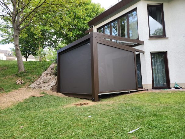 Braune Terrassenüberdachung an einem Wohnhaus, umgeben von Rasen und Bäumen.
